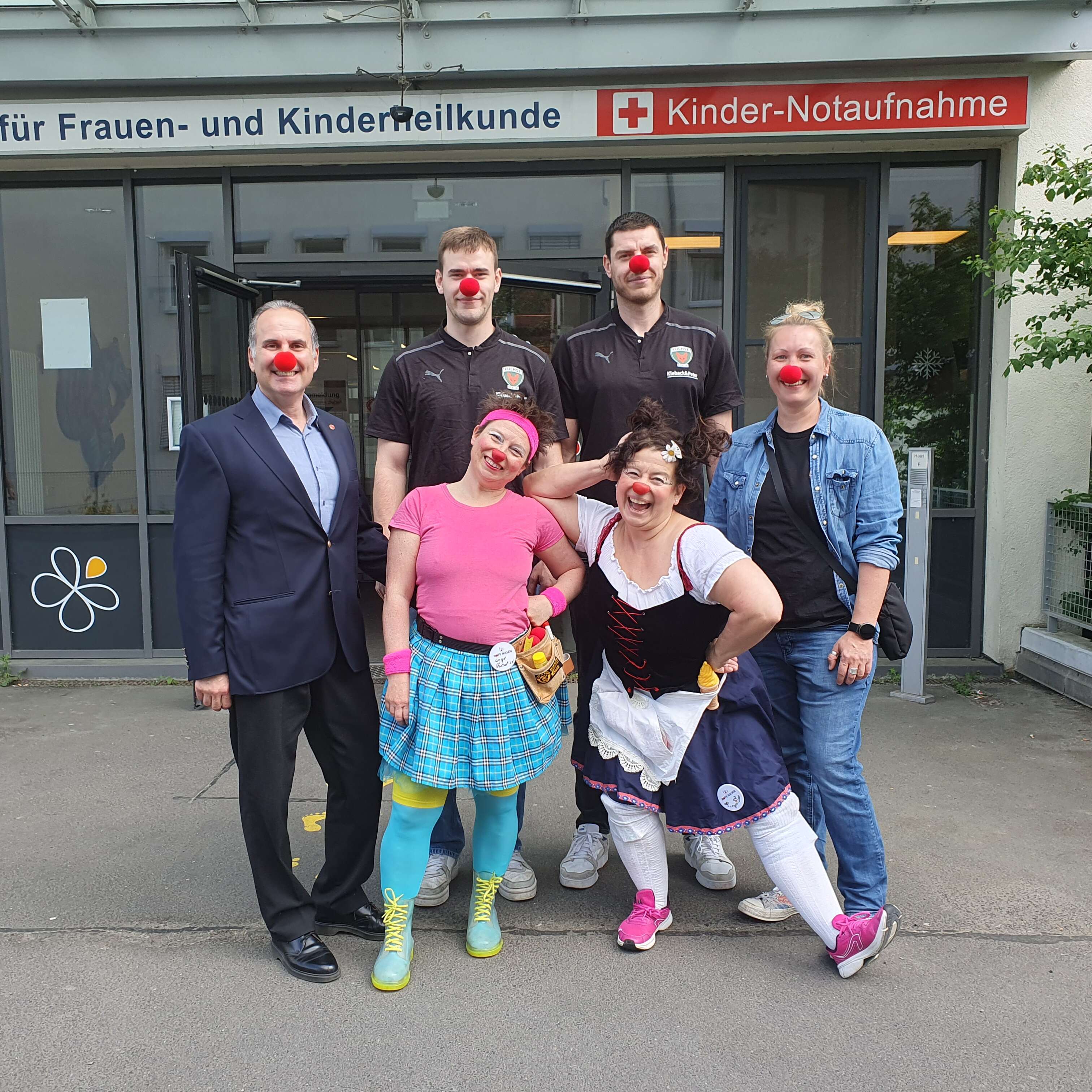 Eine Gruppe von Menschen mit roten Schaumstoffnasen steht vor dem Eingang einer Notaufnahme.