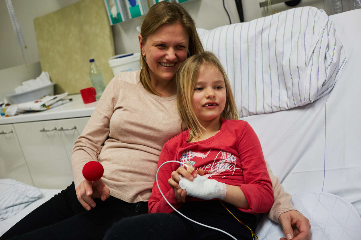 Ein Mädchen mit eine Zugang sitzt im Krankenbett zusammen mit ihrer Mutter, due eine rote Nase in der Hand hat.