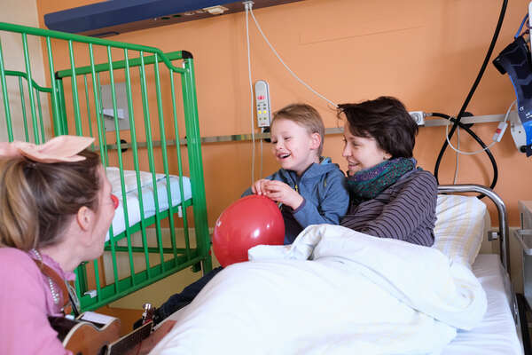 Mama und Kind sitzen im Bett und ein Clown bringt sie zum Lachen