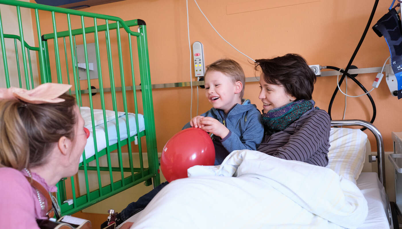Mama und Kind sitzen im Bett und ein Clown bringt sie zum Lachen