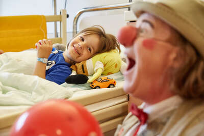 Ein jüngerer Junge freut sich über die sachen die der Klinikclown vor seinem Bett veranstaltet