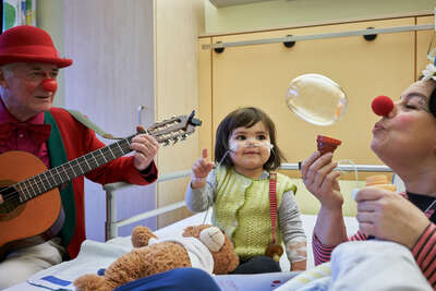 Ein kleines Mädchen schaut aus ihrem Krankenhausbättchen mit goßen kulleraugen einer Seifenblase von einem Klinikclown hinterher