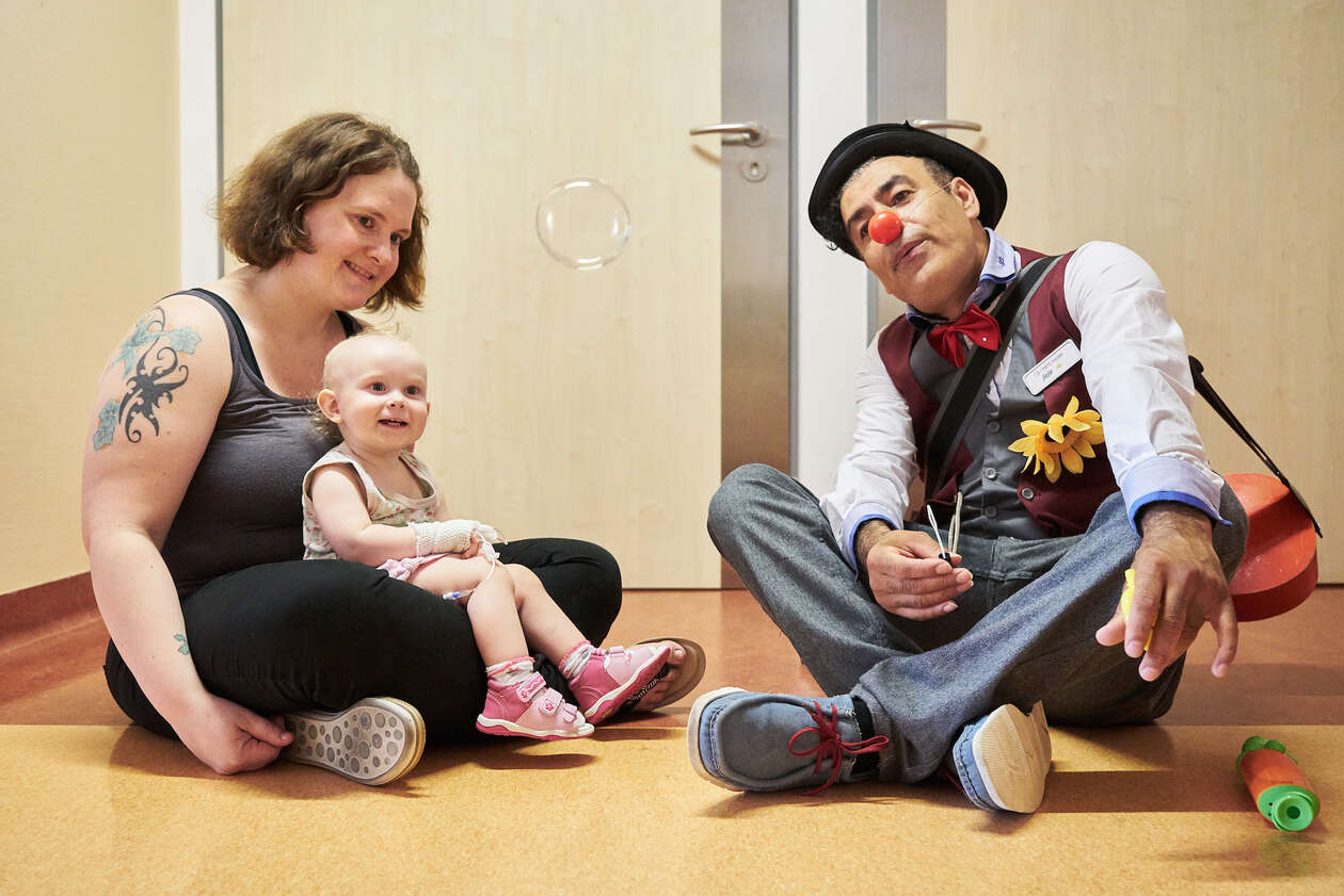Ein Clown sietzt neben einem kleinem Kind was auf dem Schoß ihrer Mutter sietzt