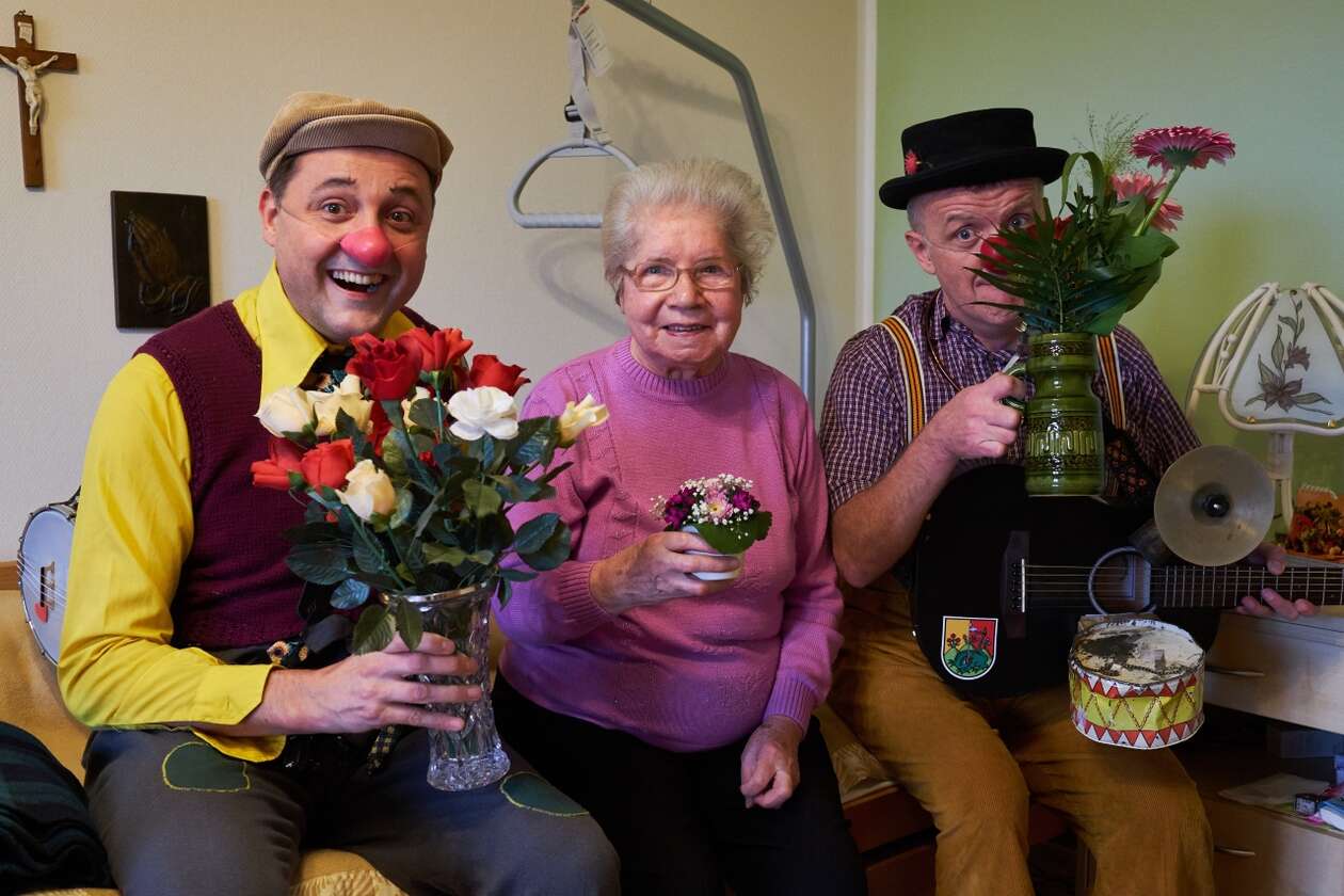 Zwei Clowns sietzen rechts und links von einer älteren Dame und sie halten alle Blumen in der Hand