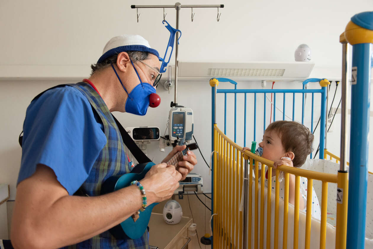 ROTE NASEN Klinikclown Dr. Meerjungblau alias Sören Kaspersinski kniet vor dem gelben Gitterbett eines Jungen in OP Hemdchen. 
