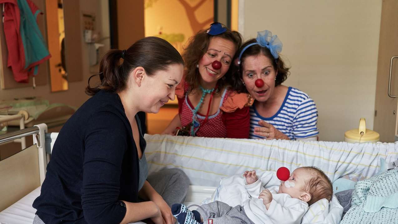 Zwei Clowns und eine junge Mutter schauen auf ein Baby mit einer roten Nasen im Krankenbett herab