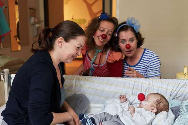 Zwei Clowns und eine junge Mutter schauen auf ein Baby mit einer roten Nasen im Krankenbett herab