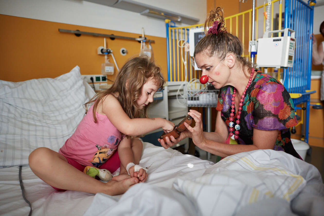 ein weiblicher Klinikclown zeigt einem klienne Mädchen im Krankenbett eine kleine Dose