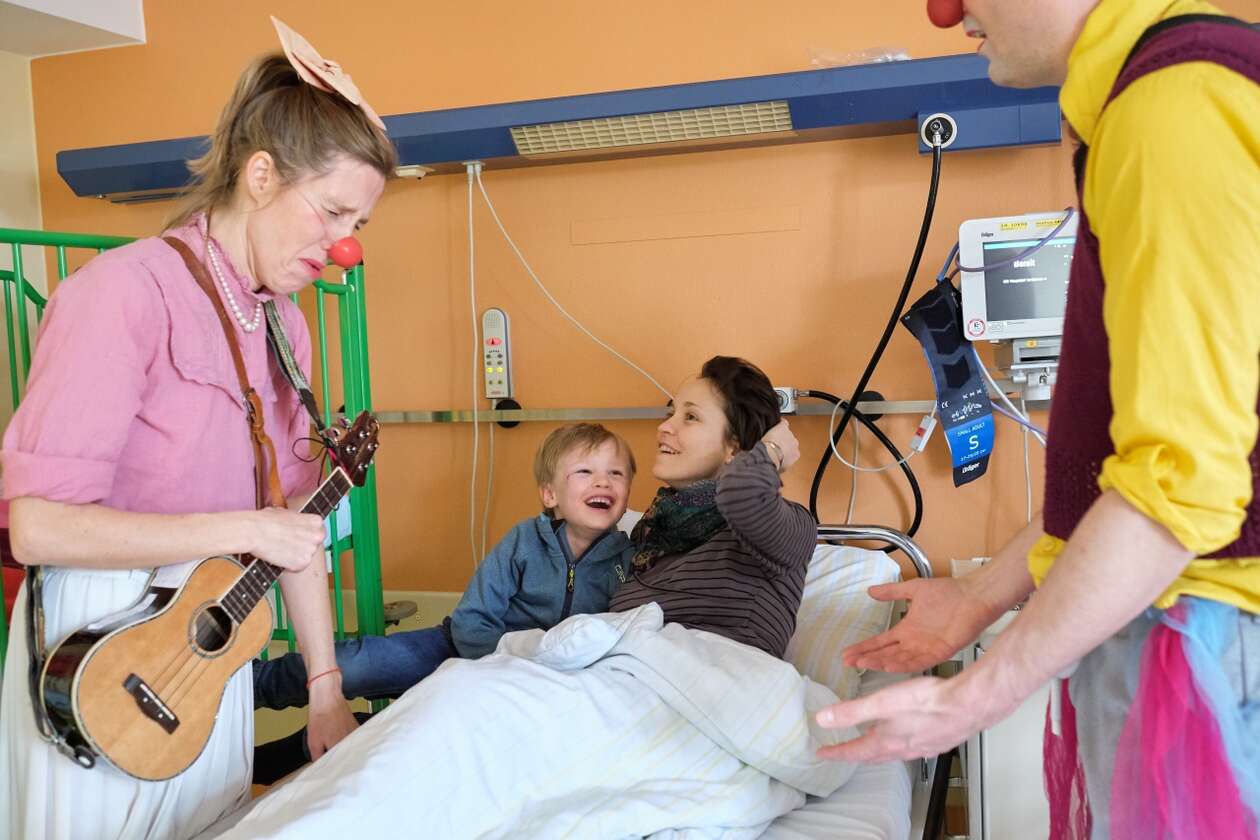 Zwei Klinikclowns stehen rechts und links von einem jüngeren Jungen und seiner Mutter am Krankenbett