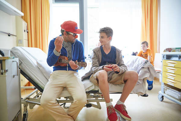 Ein Klinikclown sitzt mit einem Jugendlichen auf seinem Krankenbett