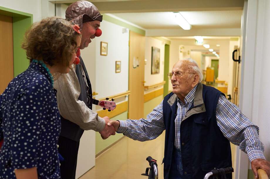 Auf dem Flur einer Pflegeeinrichtung geben zwei lachende Krankenhausclowns mit roter Nase einem Pflegebedürftigen die Hand. 