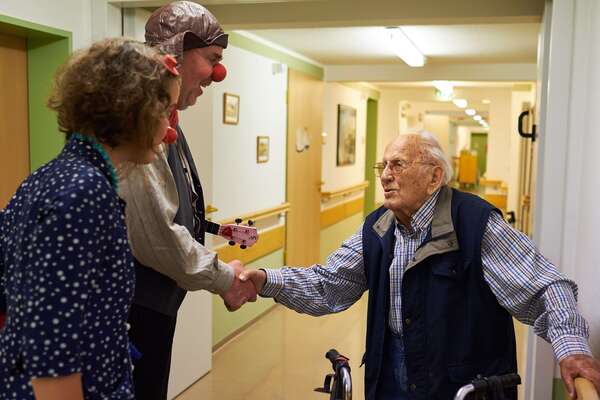 Auf dem Flur einer Pflegeeinrichtung geben zwei lachende Krankenhausclowns mit roter Nase einem Pflegebedürftigen die Hand. 