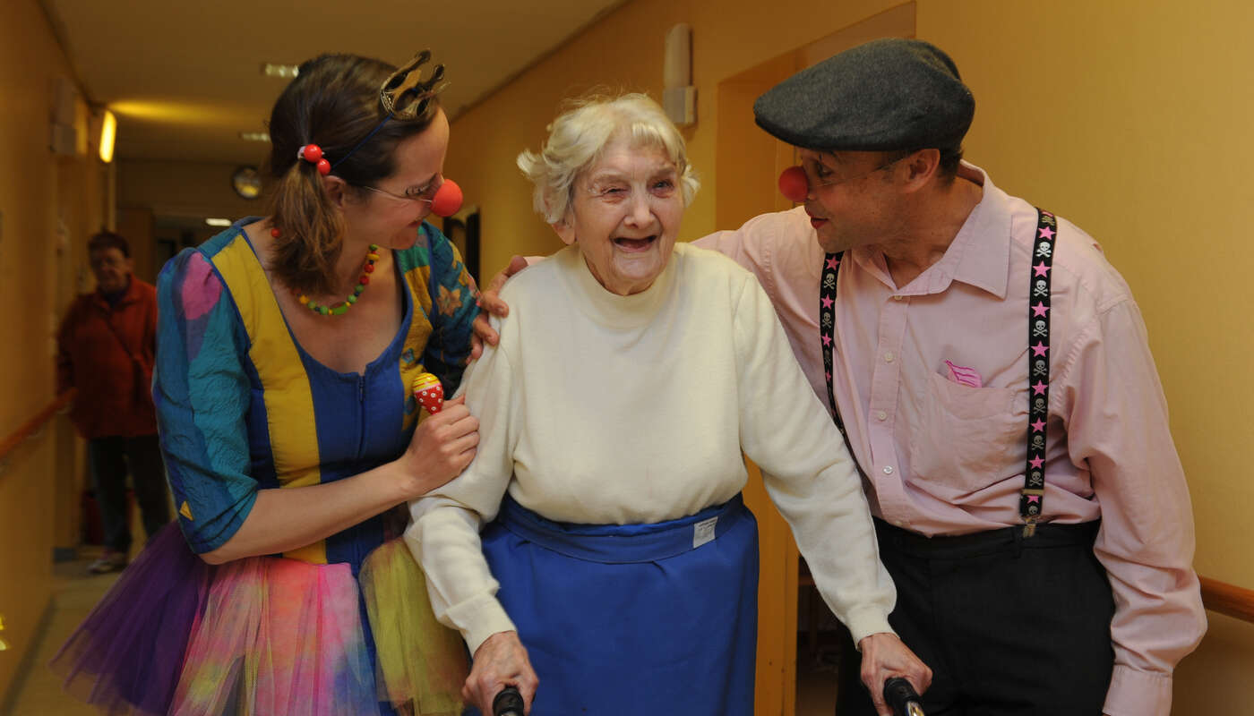 Zwei Clowns begleiten eine ältere Dame mit Rollator, die herzlich lacht