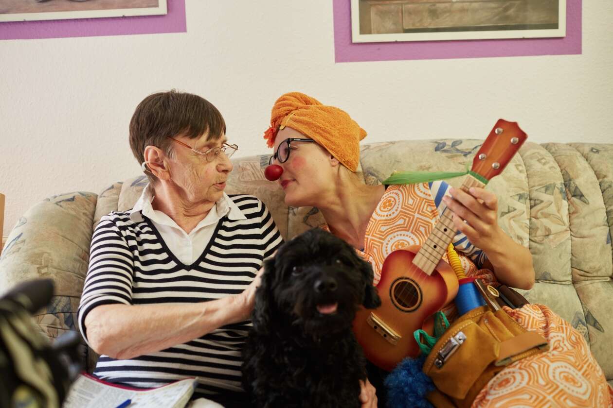 Ein Clown spielt mit ihrem Hund zusammen auf dem Sofa einer älteren Dame etwas auf ihrer Ukulele vor 