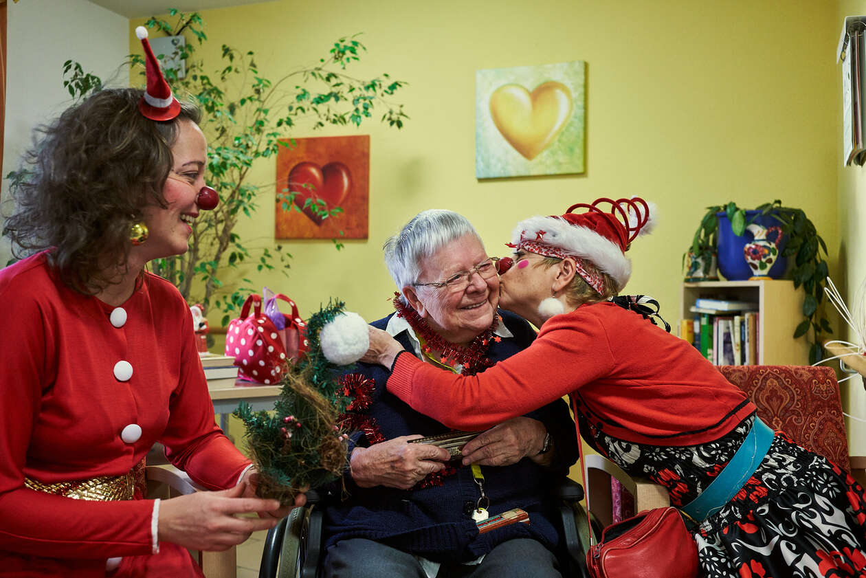 zwei weihnachtlich gekleidete Clowns sitzen neben einer älteren Dame, die Mundharmonika spielt und singen