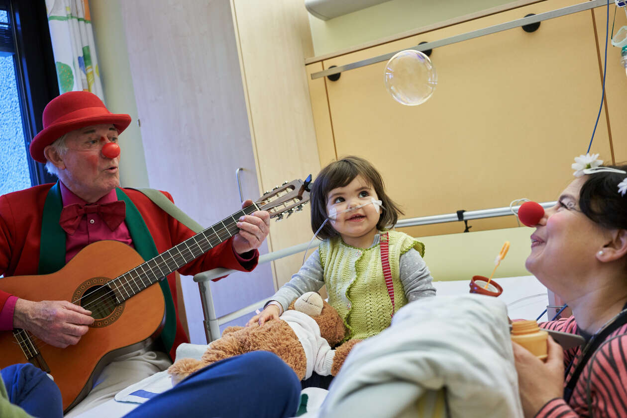 Ein kleines Mädchen schaut aus ihrem Krankenhausbättchen mit goßen kulleraugen einer Seifenblase von einem Klinikclown hinterher 