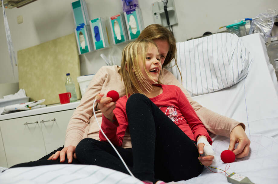 Ein Mädchen sietzt in den armen ihrer Mutter und bekommt von einem Klinikclown ein rote Nase zu geschoben 