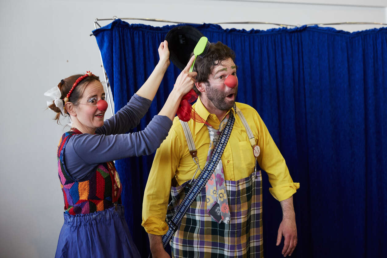 ein weiblicher Clown kämmt einem männlichen Clowns die haar mit einer überdimensionalen Zahnbürste