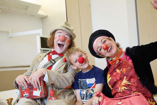 Zwei Klinikclowns haben sich neben einen Jungen auf sein Krankenhausbett gesetzt und haben ihm eine rote Nase aufgesetzt 