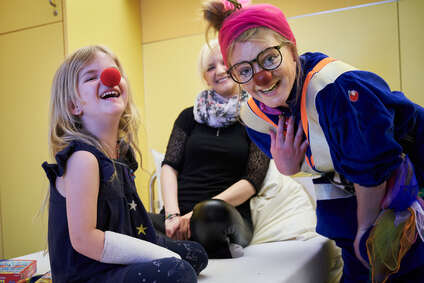 Ein blondes Mädchen mit roter Nase und ein Clown schauen in die Kamera und lachen.