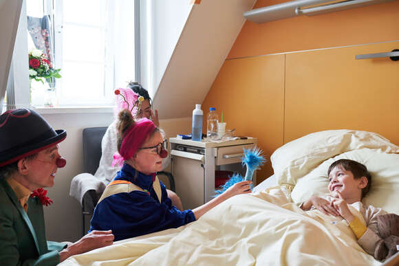 Kranker Junge liegt im Bett und wird von Clowns bespaßt.