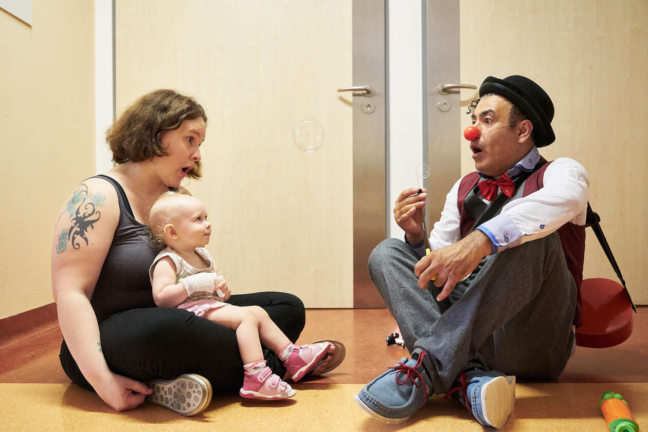 Ein Clown sietzt neben einem kleinem Kind was auf dem Schoß ihrer Mutter sietzt