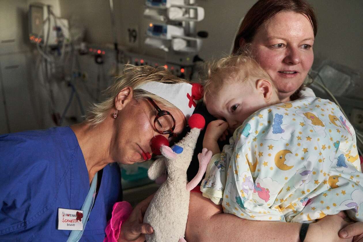 ROTE NASEN Clown Brischitt und ihre Kuscheltierratte Gustav kuschenl mit dem kleinen Luca, der am Herzen operiert werden soll. Seine Mutter schaut besorgt.