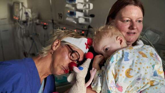 ROTE NASEN Clown Brischitt und ihre Kuscheltierratte Gustav kuschenl mit dem kleinen Luca, der am Herzen operiert werden soll. Seine Mutter schaut besorgt.