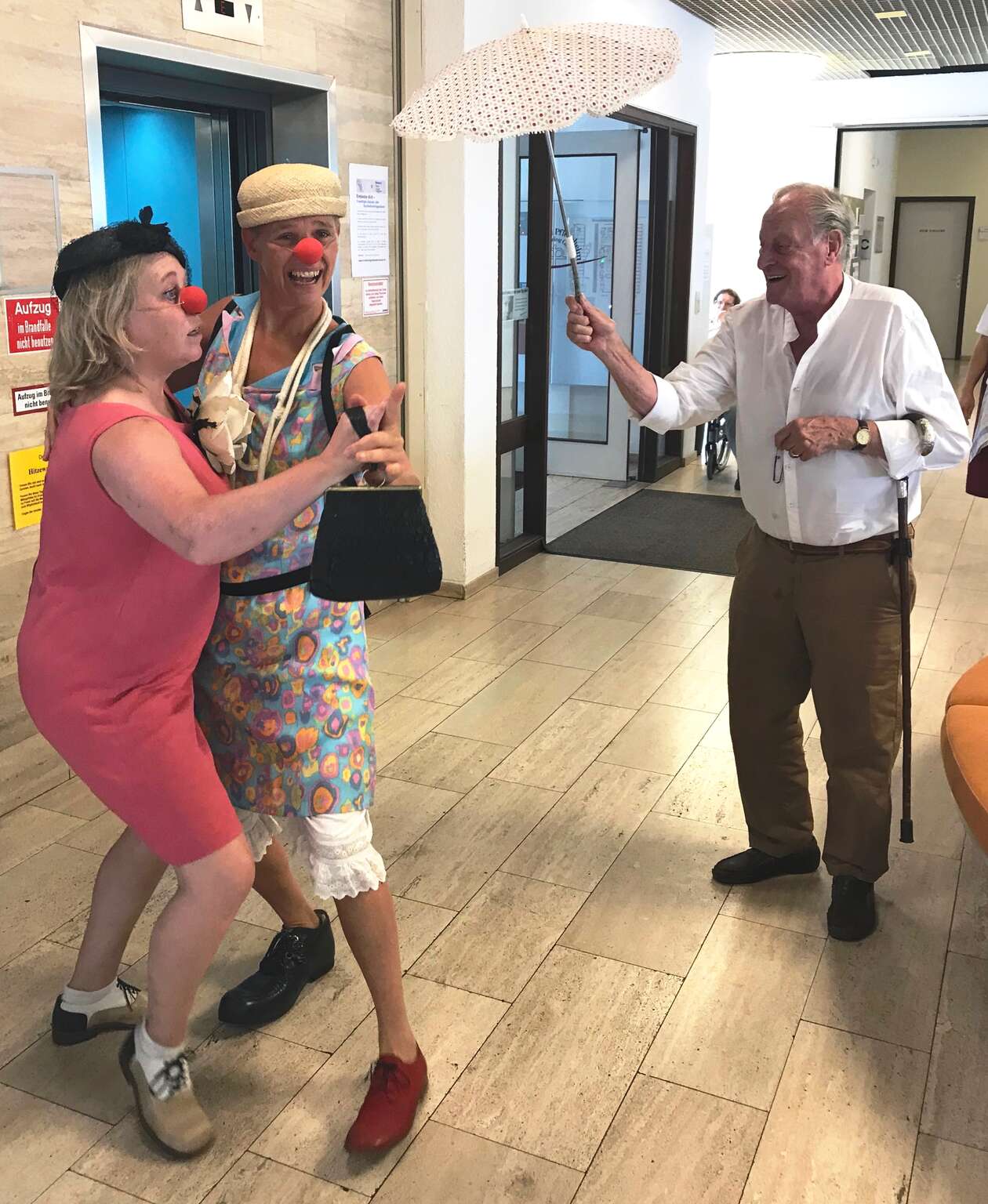 Zwei Clowns tanzen vor einem älteren Herren im Flur vom Pflegeheim 