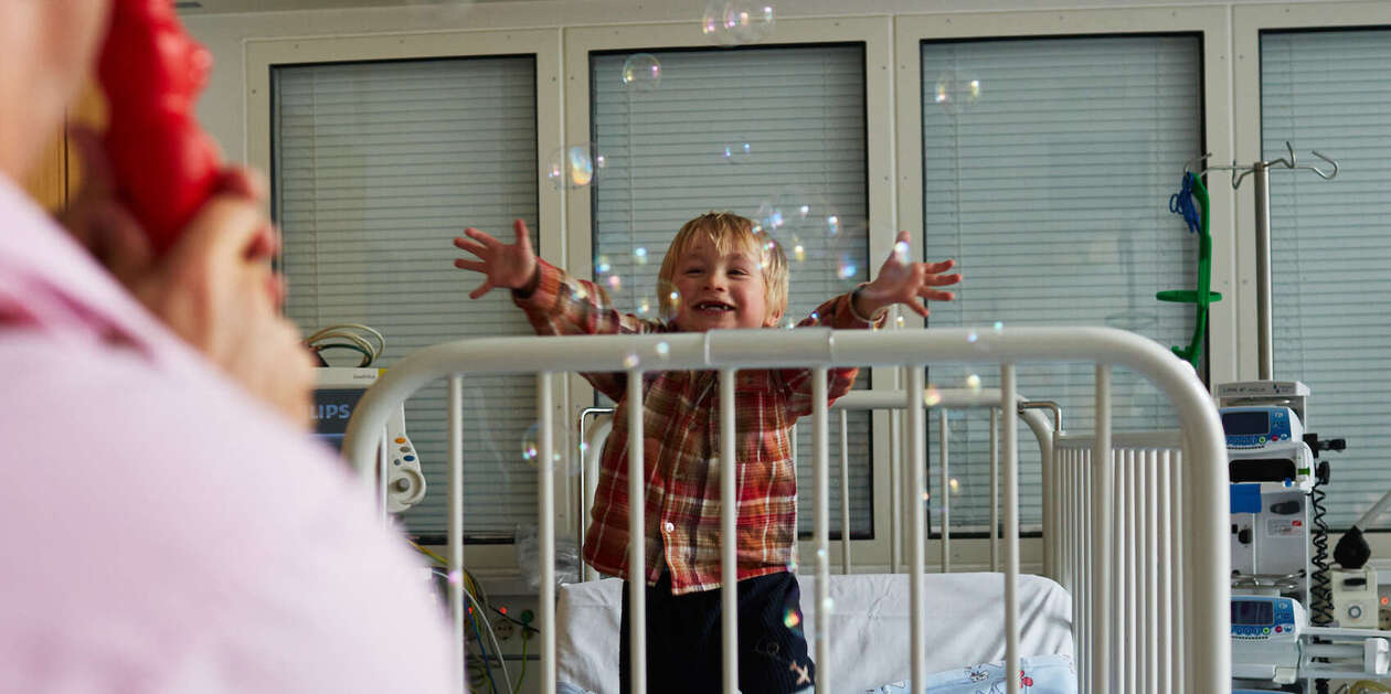 Ein Clown pustet ganz viele kleine Seifenblasen in ein Krankenzimmer von einem kleinem Jungen, der versucht nach ihnen zu greifen 