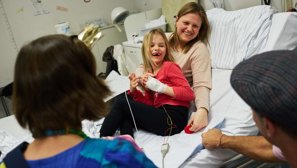Ein Mädchen sietzt in den armen ihrer Mutter und bekommt von einem Klinikclown ein rote Nase zu geschoben 