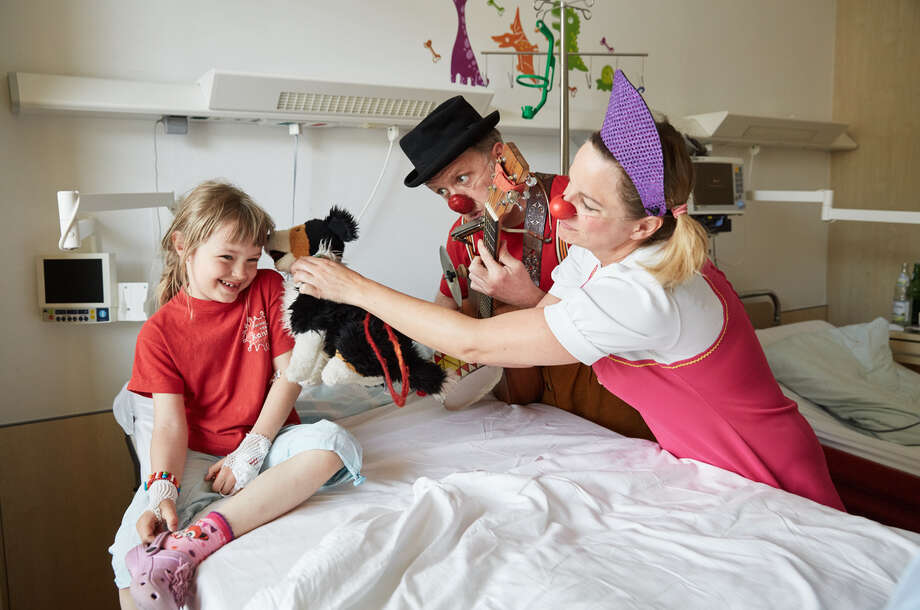 Ein in rot gekleidetes Mädchen sitzt im Krankenbett und zwei Clowns machen Musik und spielen mit ihrem Hunde-Kuscheltier