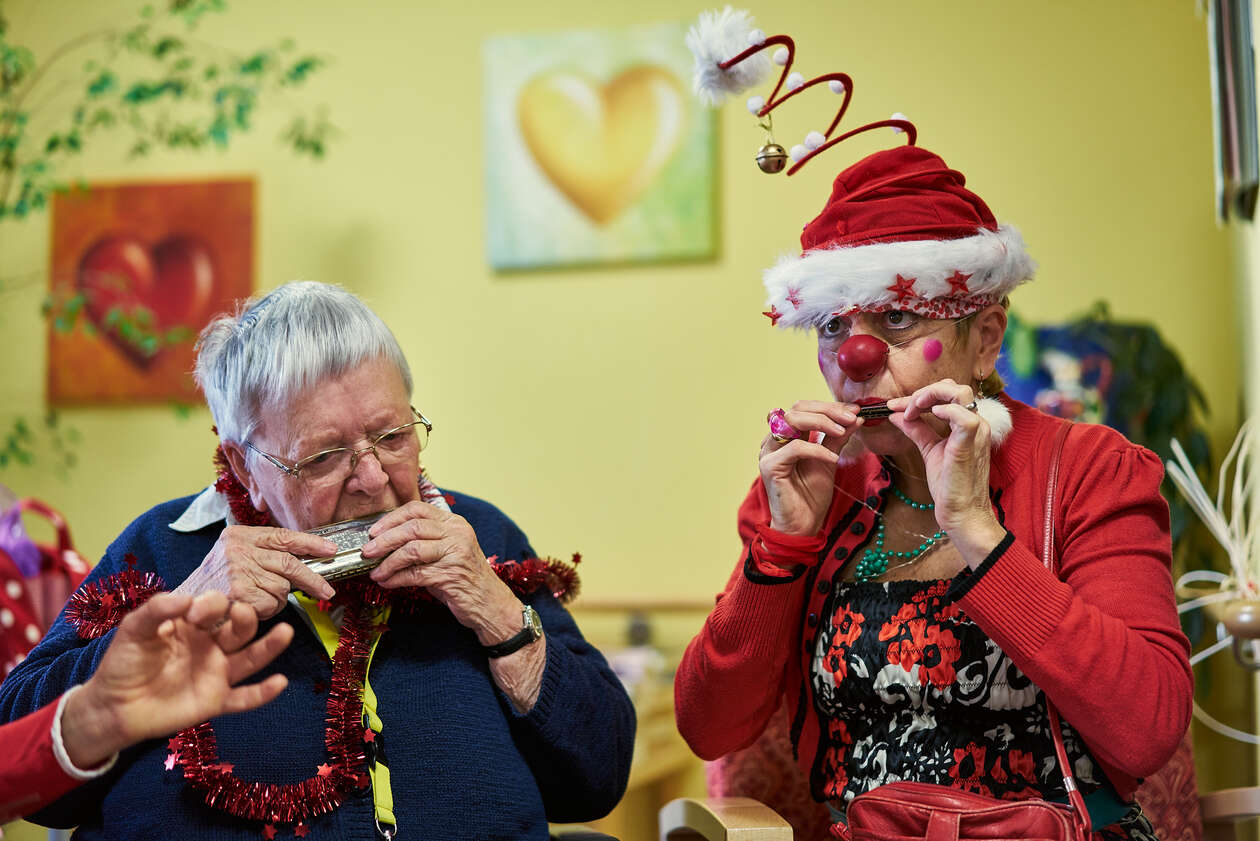 ein weihnachtlich gekleideter Clown sitzt neben einer älteren Dame und beide spielen ihre Mundharmonika
