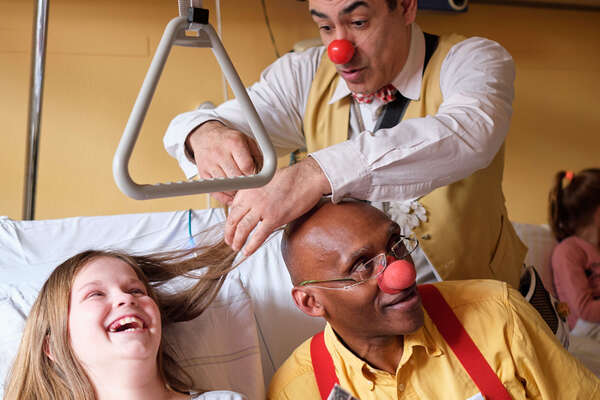 Zwei männlcihe Clowns stehen an eine Krankenbett, wo ein Teenager Mädchen liegt und herzhaft lacht