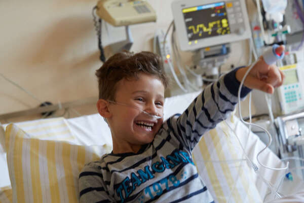 Ein Junge zeigt von seinem Krankenbett aus mit seinem zeigefinger lachet auf die Klinikclowns