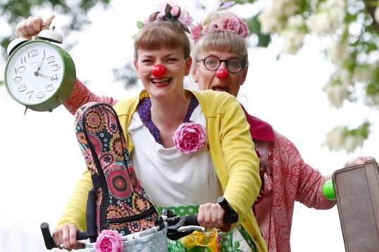 Zwei Clowns witzen auf einem Fahrrad und zeigen eine große Uhr und einen Koffer.