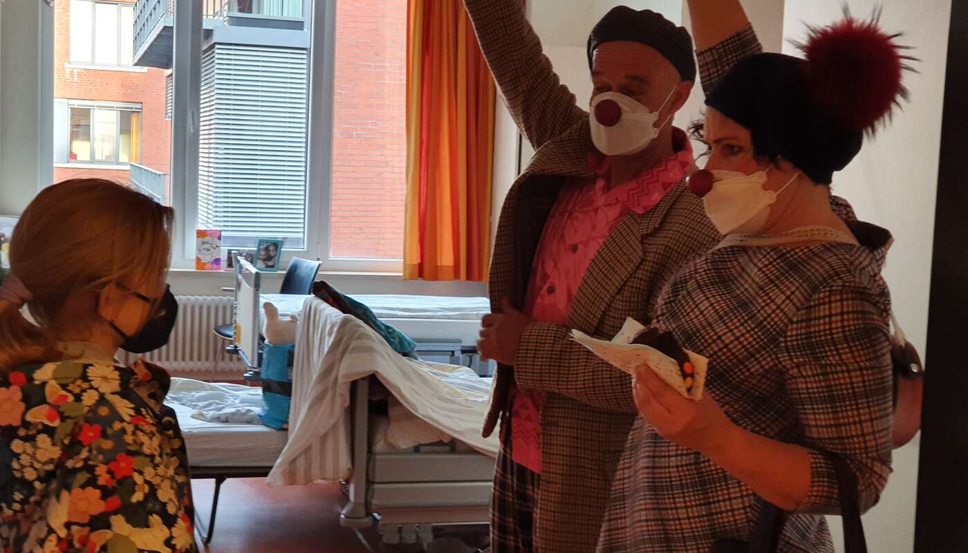 2 Clowns stehen im Zimmer einer Patientin und raten ein Ratespiel mit Kuchen in der Hand
