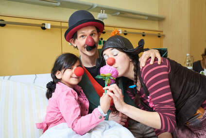 Ein Mädchen mit roter Schaumstoffnase schaut zusammen mit zwei Klinikclowns lachend in die Kamera.