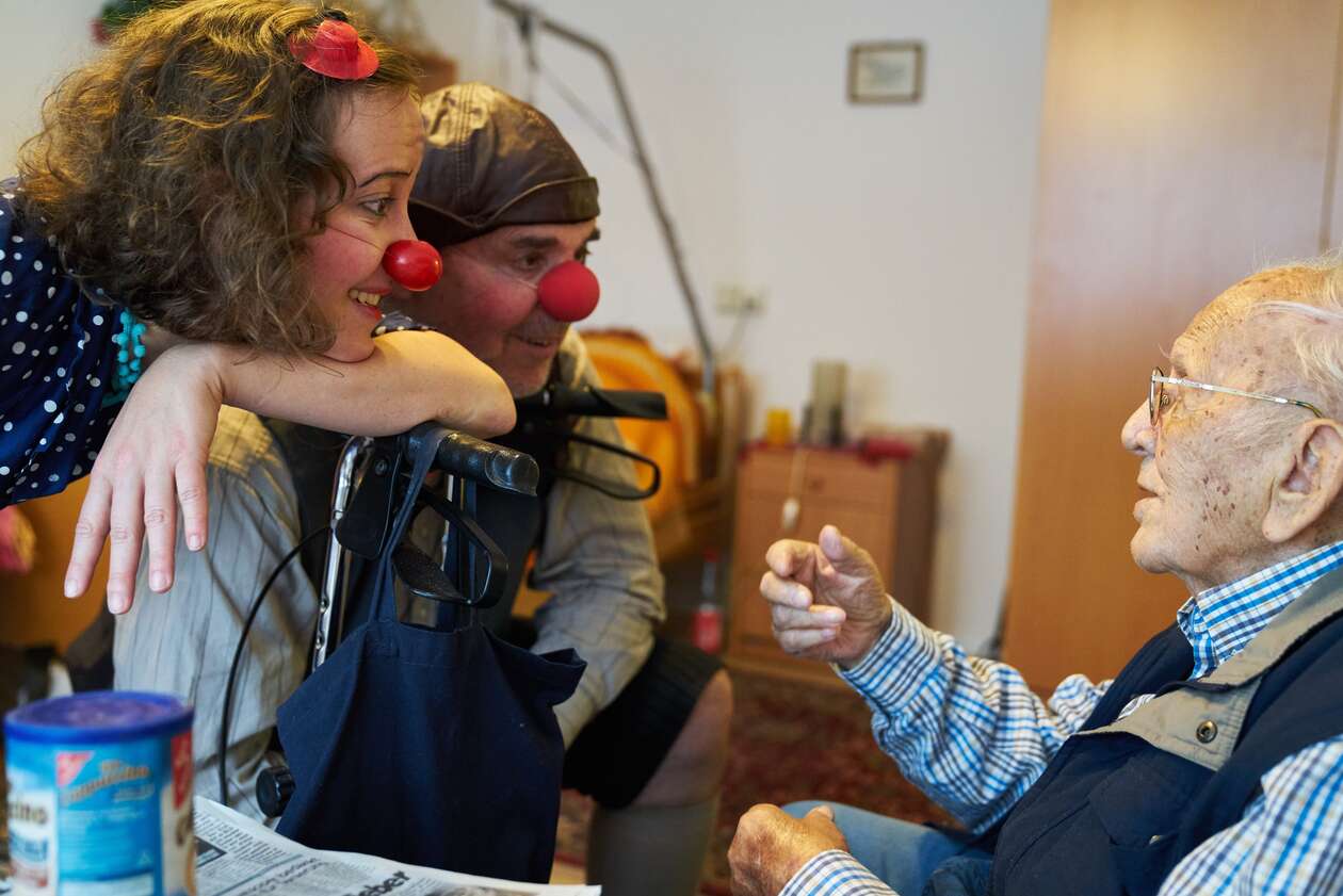 Zwei Klinikclowns mit roter Nase hören lächelnd einem pflegebedürftigen Mann zu