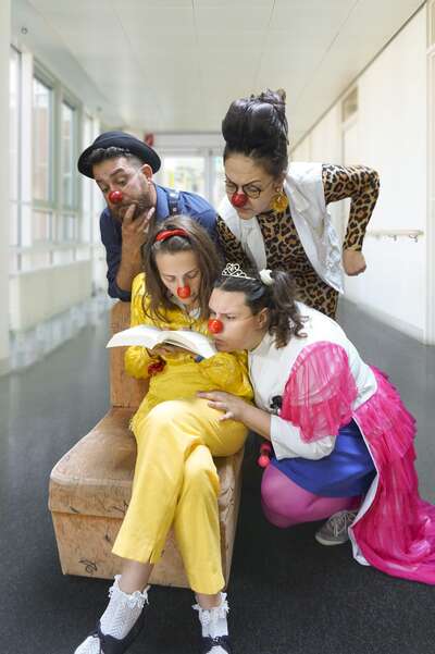 Eine Clownin sietzt auf einem hocker und schaut in ein Buch. Drei andere Clowns stehen hinter ihr und schauen ihr über die schulter