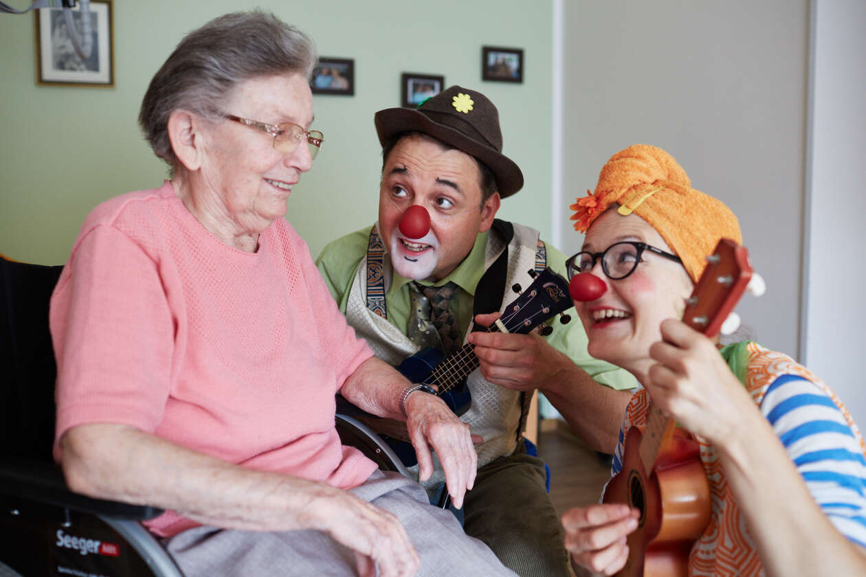 Zwei Clowns hocken neben einer älteren Dame im Rollstuhl un singen ihr was vor