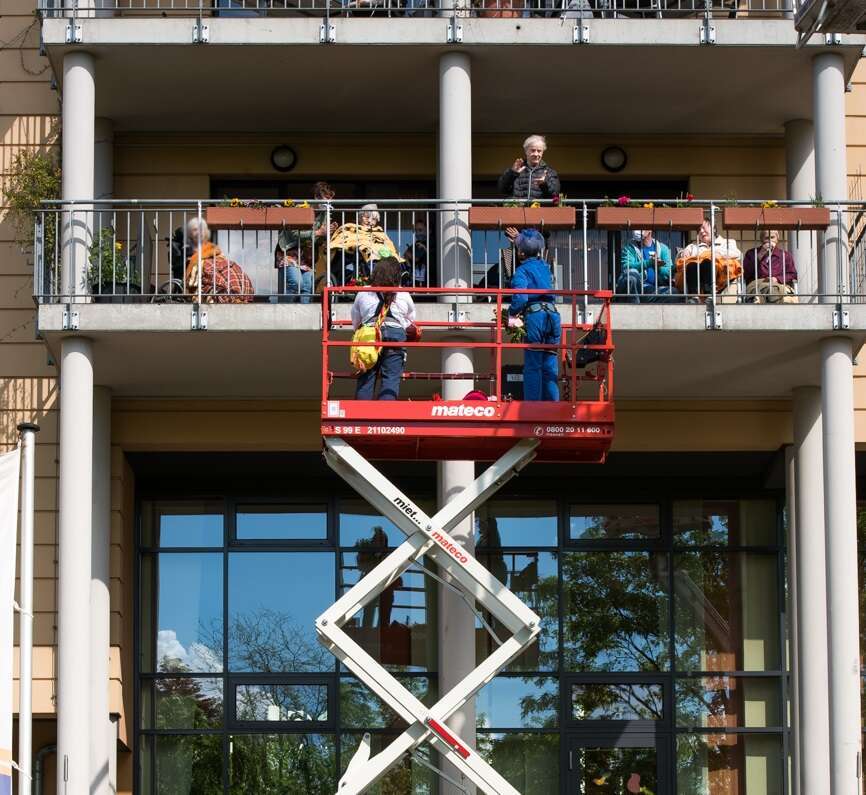 Clowns stehen auf einer Hebebühne und spielen für ältere Menschen im Seniorenheim, die auf dem Balkon sitzen.