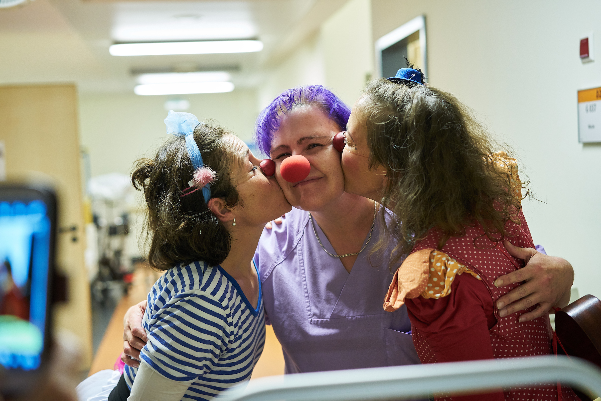 Zwei Klinikclowns geben einer Pflegekraft von jeder seite einen kuss auf die wange 