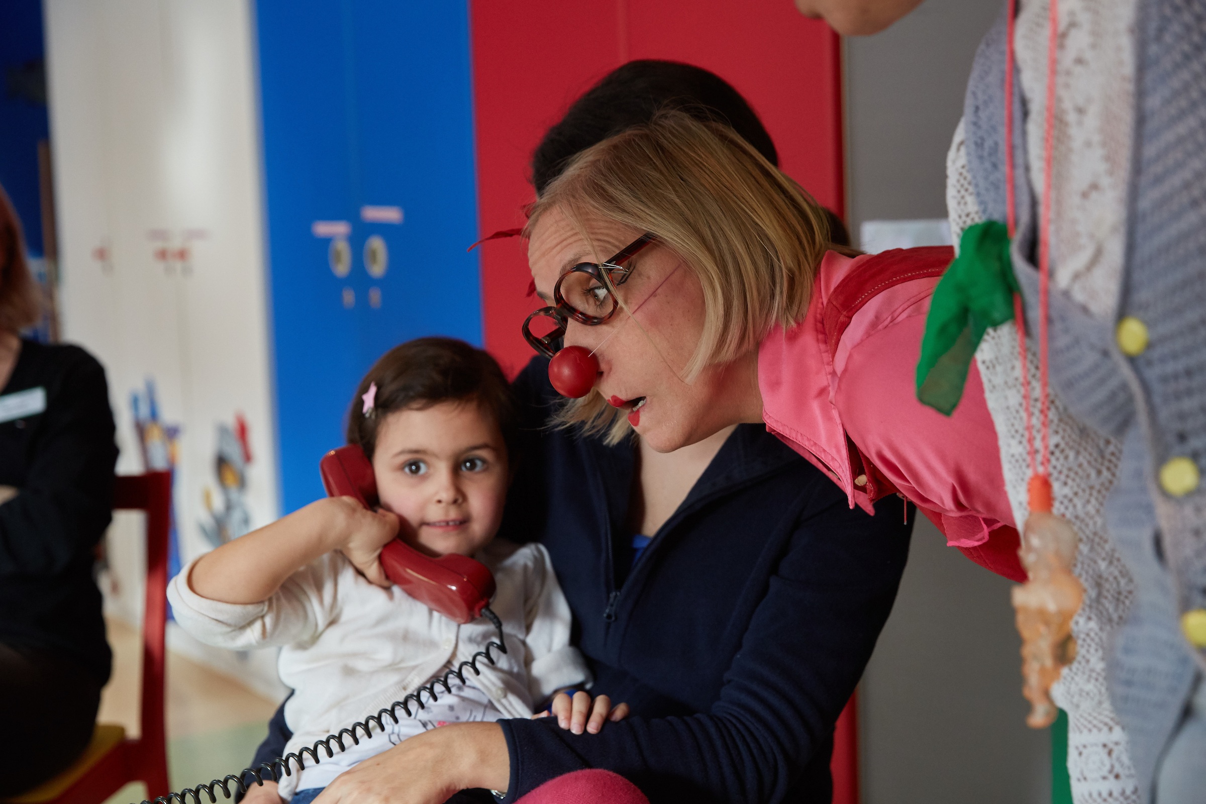 Eine Clownvisite: Ein Mädchen im Krankenhaus hat einen Telefonhörer in der Hand, ein weiblicher Klinikclown lauscht