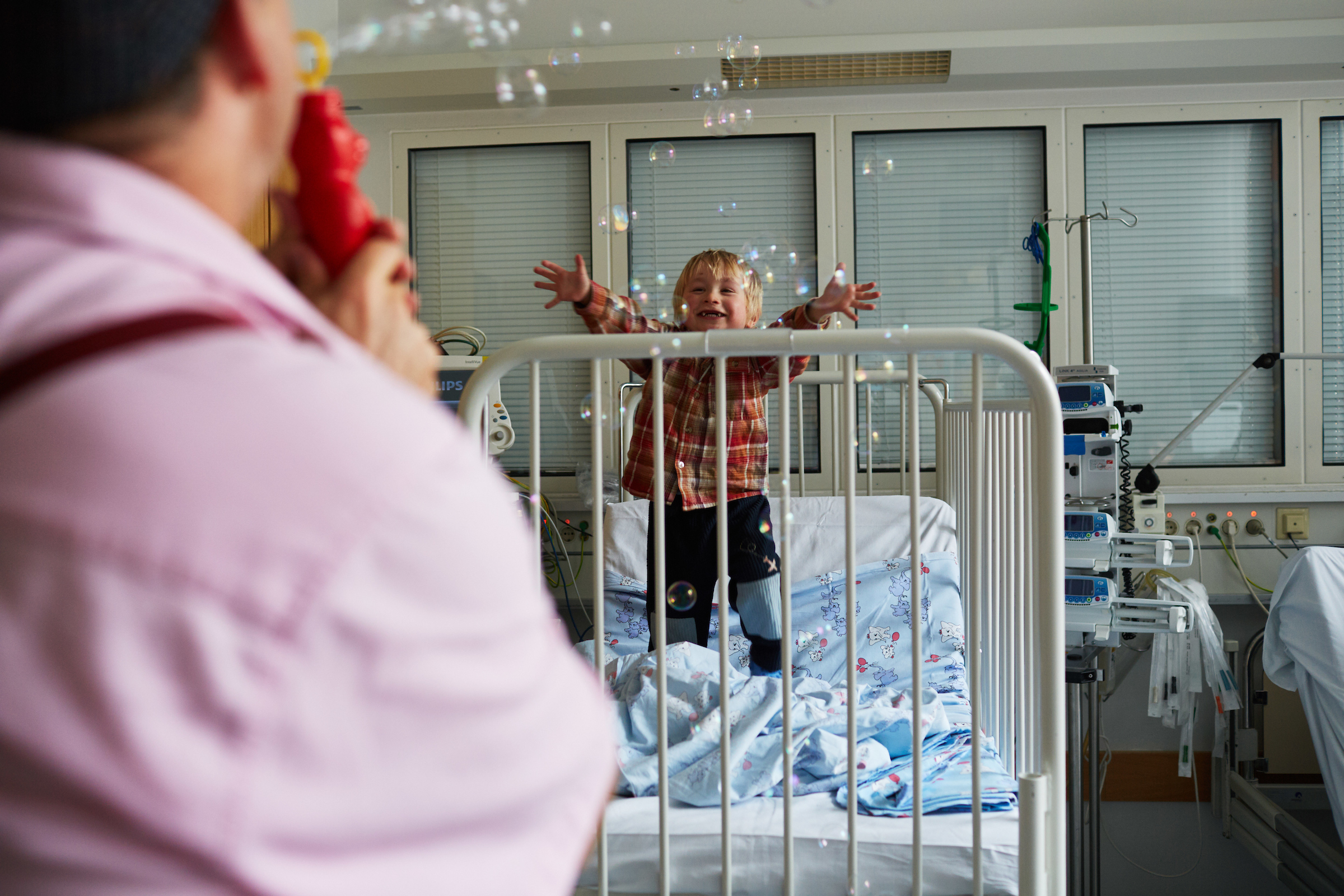Ein Clown pustet ganz viele kleine Seifenblasen in ein Krankenzimmer von einem kleinem Jungen, der versucht nach ihnen zu greifen
