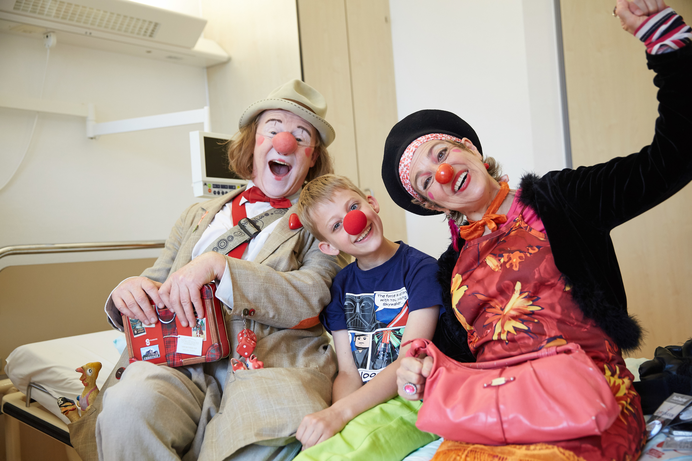 Zwei Klinikclowns haben sich neben einen Jungen auf sein Krankenhausbett gesetzt und haben ihm eine rote Nase aufgesetzt