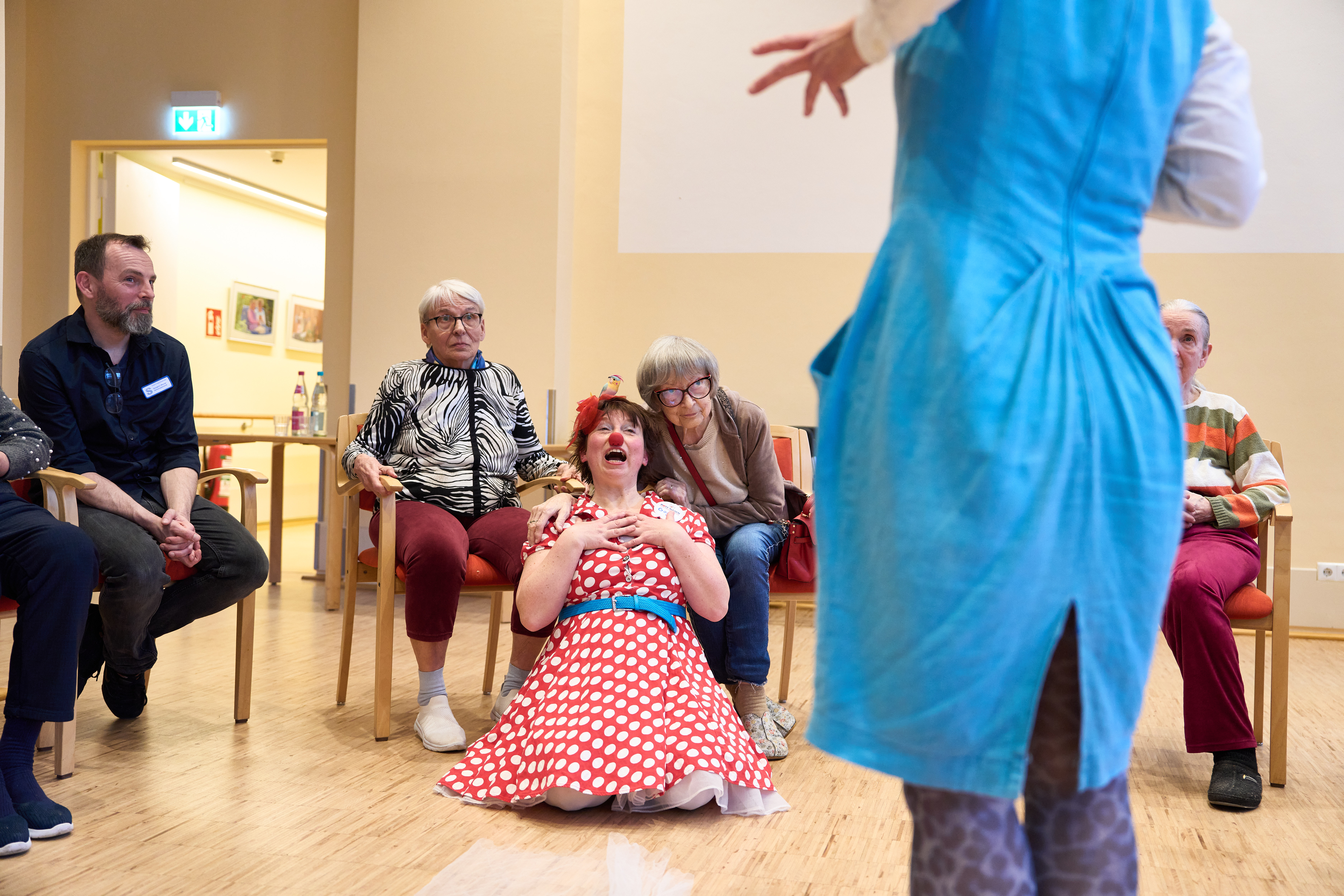 Ein Clowns sitzt auf dem Boden vor zwei älteren Damen auf Stühlen