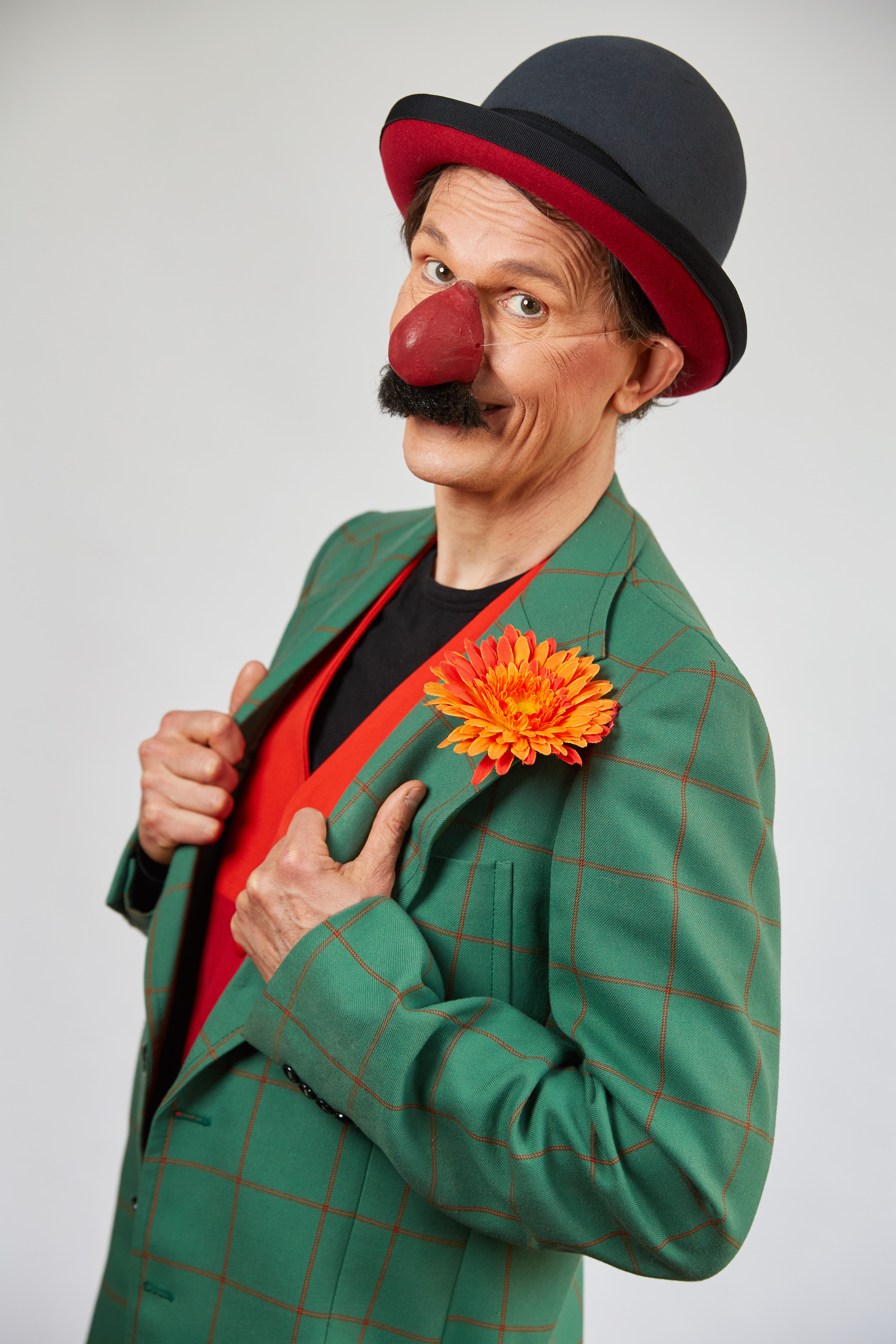 Porträtfoto von Clown nartusch mit einerm grünen Blazer, einem Hut und einer orangefarbenen Blume