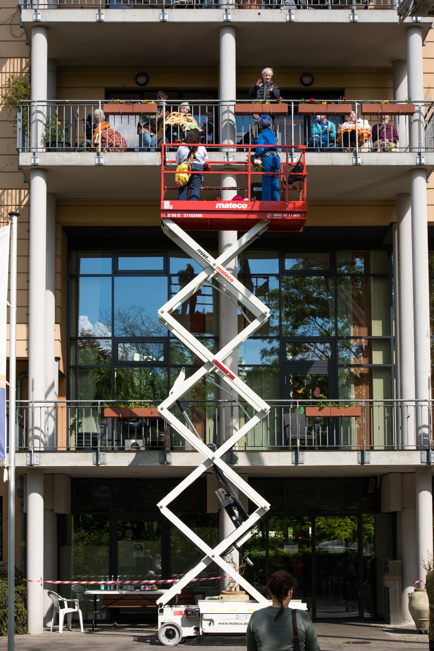 Clowns stehen auf einer Hebebühne und spielen für ältere Menschen im Seniorenheim, die auf dem Balkon sitzen.