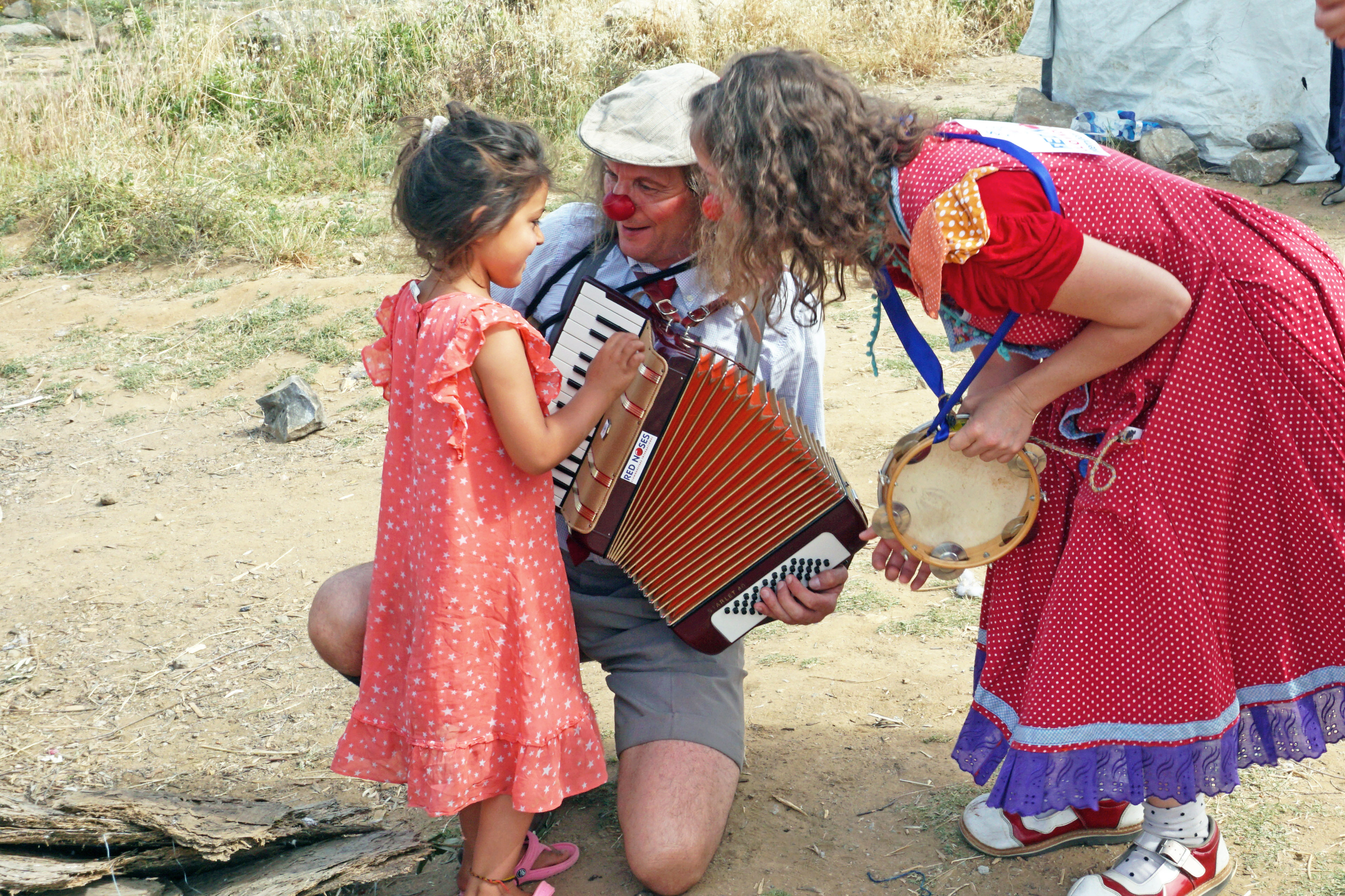 ROTE NASEN Clown Luciana im Flüchtlingscamp in Lesbos beugt sich mit roter Nase zu einem geflüchteten Mädchen hinunter. Ein anderer Klinikclown hat ein Akkordeon in der Hand. Im Rahmen des Programms Emergency Smile.